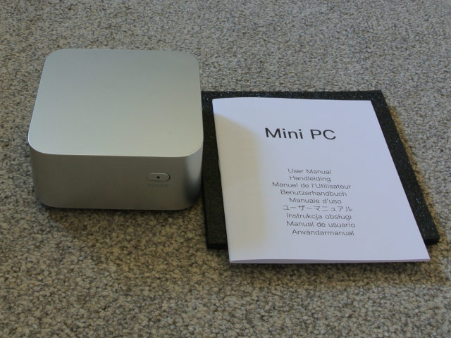 An anterosuperior oblique view of the device, which is a compressed silvery cube with rounded corners. There is a single power button on the front panel. The surface area of the device is smaller than the adjacent instruction manual